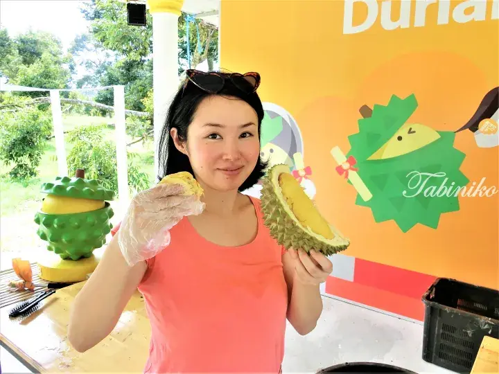 Woman holding fresh durian at a Malaysia fruit farm with a Durian-themed wall and green mascot.