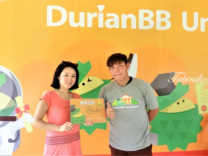 A couple poses with a DurianBB University graduation certificate in front of a yellow promotional wall.
