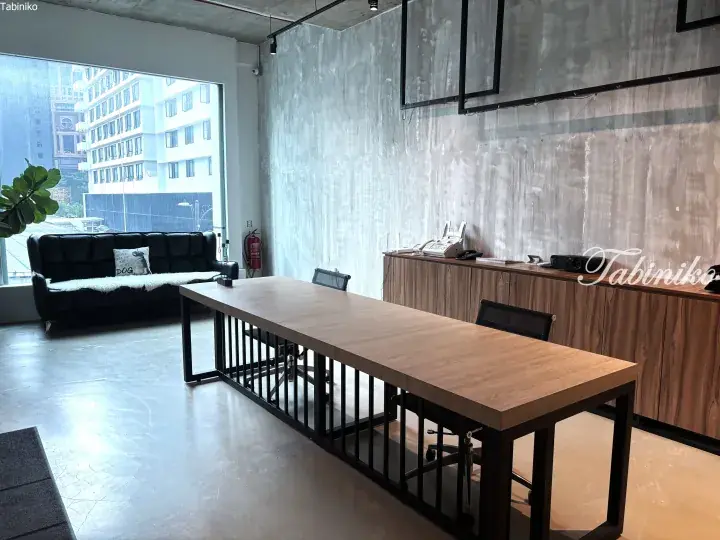 Modern industrial office space featuring a long wooden table, concrete walls, and a black leather sofa by a large window.