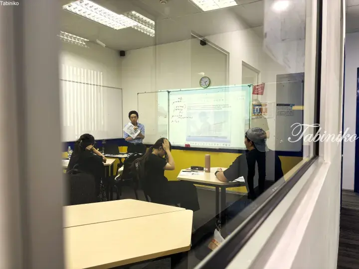 Teacher instructing students in a bright classroom with whiteboards and a projector screen during an English lesson.