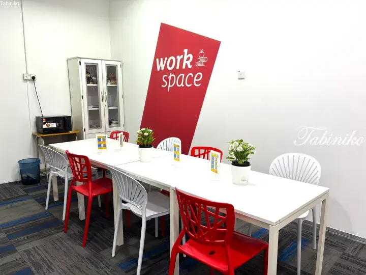 Bright workspace with red and white chairs around a table, featuring a red wall sign and potted plants.