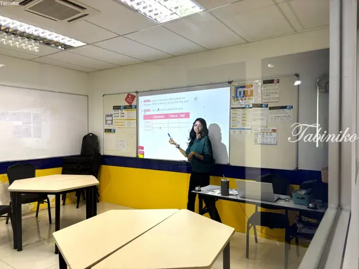 Female teacher instructing students in a bright classroom with yellow and blue walls, pointing at a whiteboard displaying English lesson content.