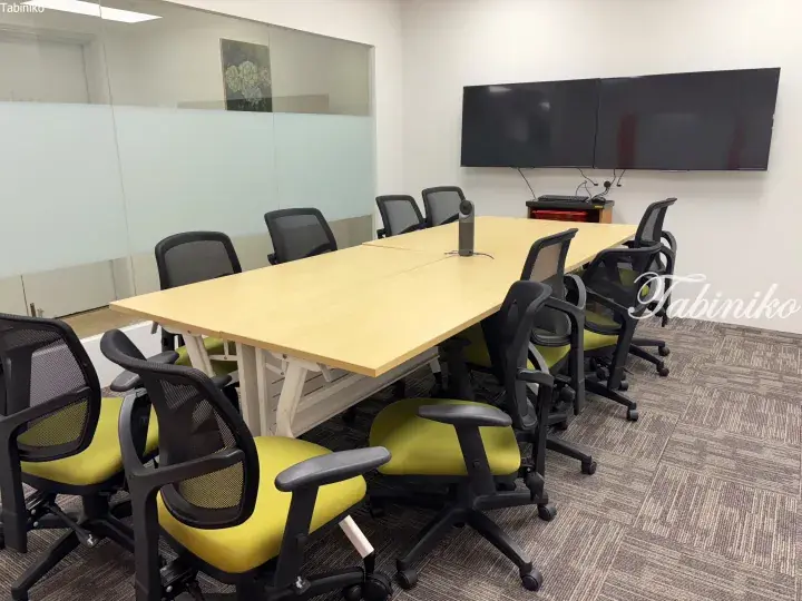 Modern meeting room with a large wooden table, green office chairs, and dual wall-mounted screens.