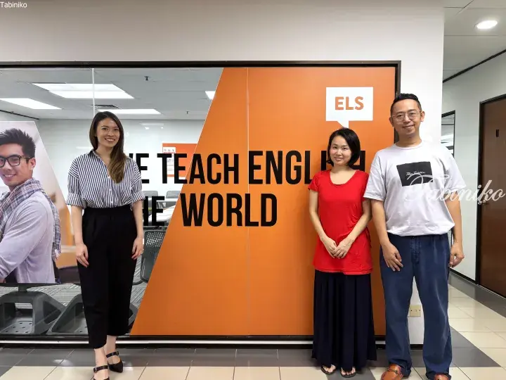 Three people standing in front of an orange wall with ELS logo and text ‘WE TEACH ENGLISH THE WORLD’.