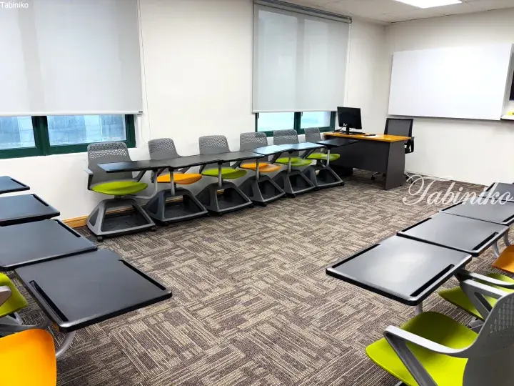 Modern classroom with grey carpet, whiteboard, and colorful student desks arranged in rows.