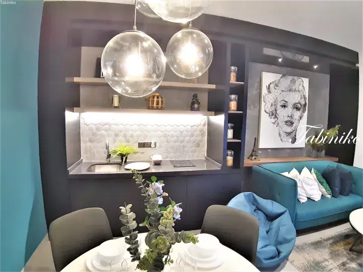 Modern studio apartment interior featuring a compact kitchenette with hexagonal tiles, a teal sofa, and a geometric Marilyn Monroe wall art.
