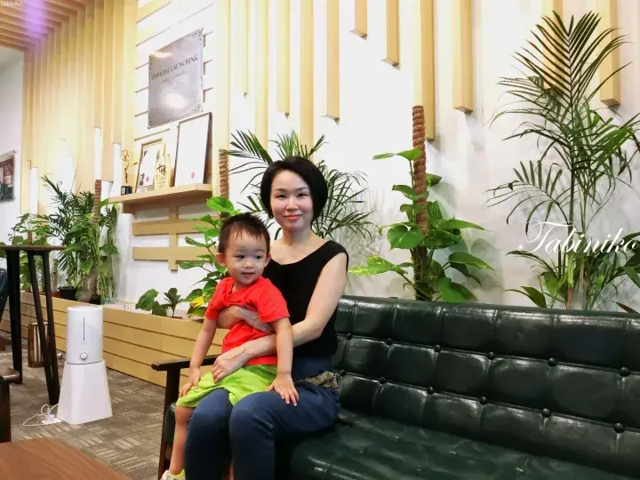 Woman and child sitting on a green leather sofa in the lobby of Embique Condotel, featuring wooden decor and plants.