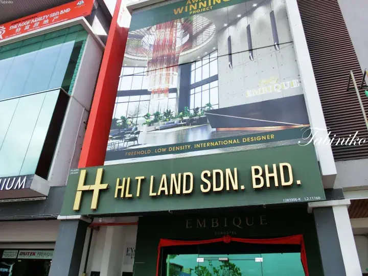 The sales gallery entrance for Embique Condotel, featuring the developer’s HLT Land SDN BHD sign and a large banner showcasing the project’s modern interior design.