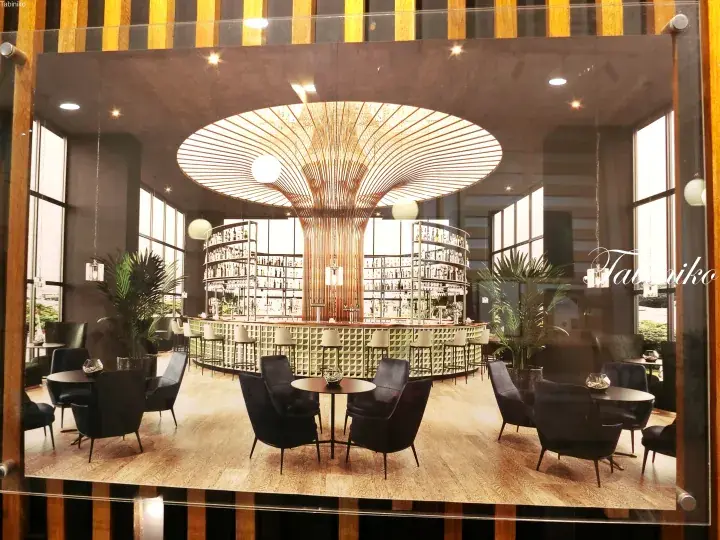 Modern bar interior with a large wooden canopy light fixture, circular shelving, and black seating.