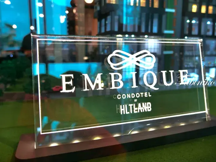 Close-up of the EMBIQUE Condotel by HLT Land logo on a clear acrylic display stand.