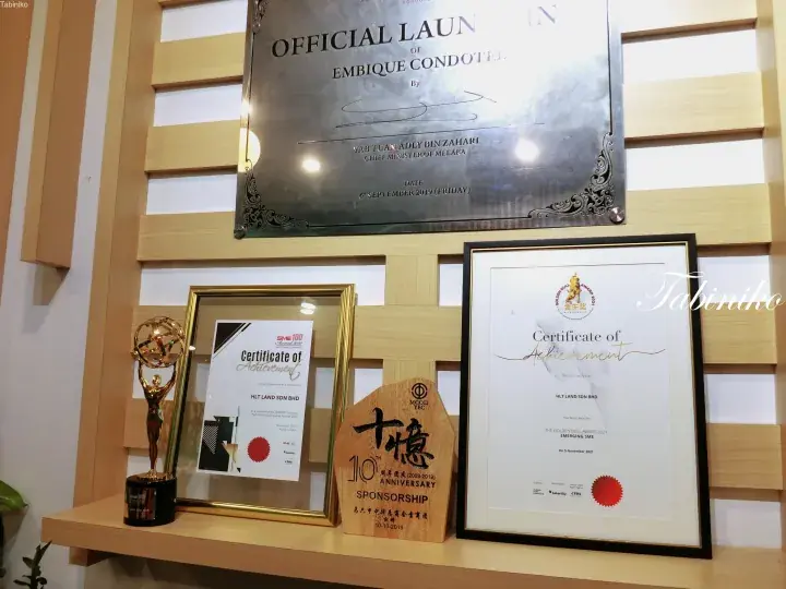 Awards and certificates displayed on a wooden shelf, including the official launch plaque for Embique Condotel.