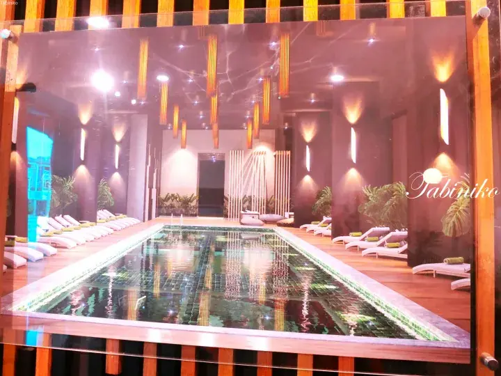 Modern indoor swimming pool with green tiles, wooden deck, lounge chairs, and elegant pendant lighting.