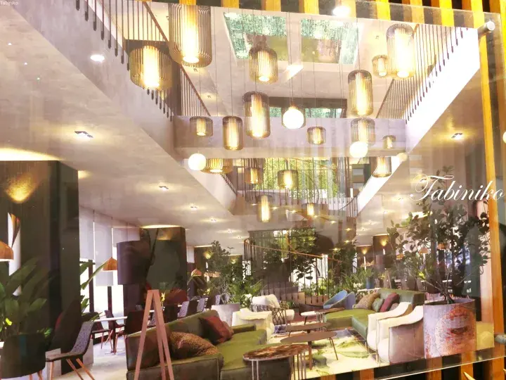 Modern hotel lobby with high ceilings, hanging pendant lights, green sofas, and indoor plants.