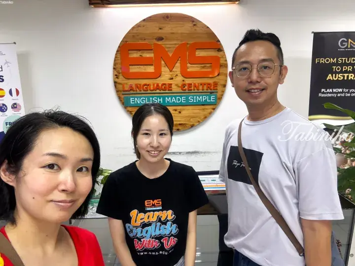 Three people smile in front of the EMS Language Centre sign in Malaysia.