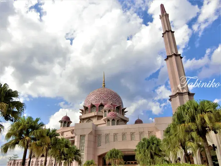 Pink-domed Putra Mosque in Putrajaya, Malaysia, featuring intricate geometric patterns and a tall minaret against a cloudy blue sky.