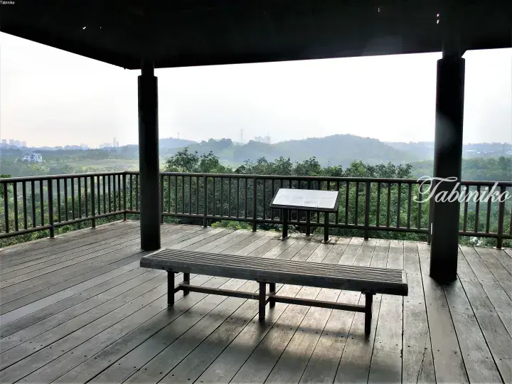 Wooden viewing deck with bench and information board overlooking lush green hills and distant city skyline.