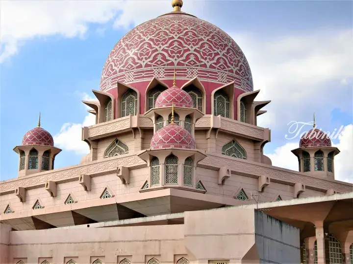 Pink domes with intricate white geometric patterns and arched windows of a mosque against a blue sky.