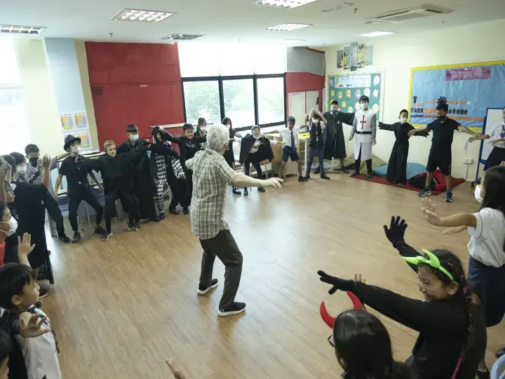 An older man leads a group of students in a classroom activity where children wear costumes like knights and witches.