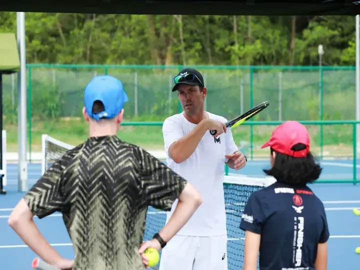 Tennis coach demonstrating a swing technique to two young students on an outdoor court.