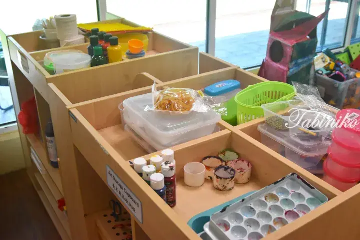 Organized wooden art storage cupboard with paint bottles, containers, and craft supplies in a bright room.