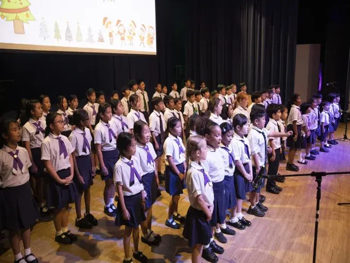 Students in uniform singing in a choir on a school stage with Christmas decorations projected on a screen.