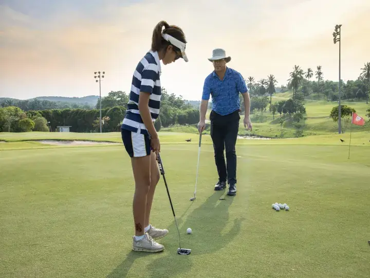 A woman practices putting on a lush green golf course while a man observes in the background.