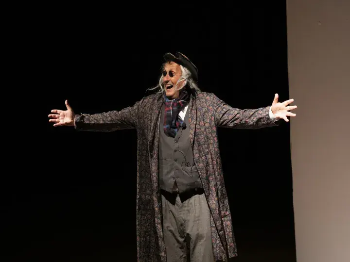 A man in a patterned coat and hat performs on stage with arms outstretched and a joyful expression.