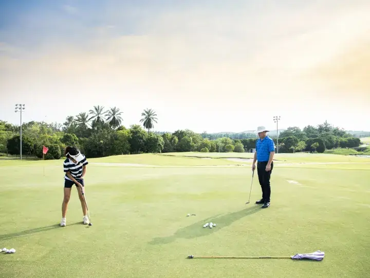 A woman putting on a golf green while a man in a blue shirt watches nearby on a sunny day.