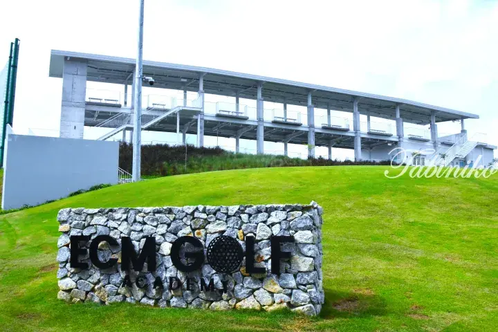ECM Golf Academy sign on a stone wall with a modern driving range pavilion on a grassy hill.