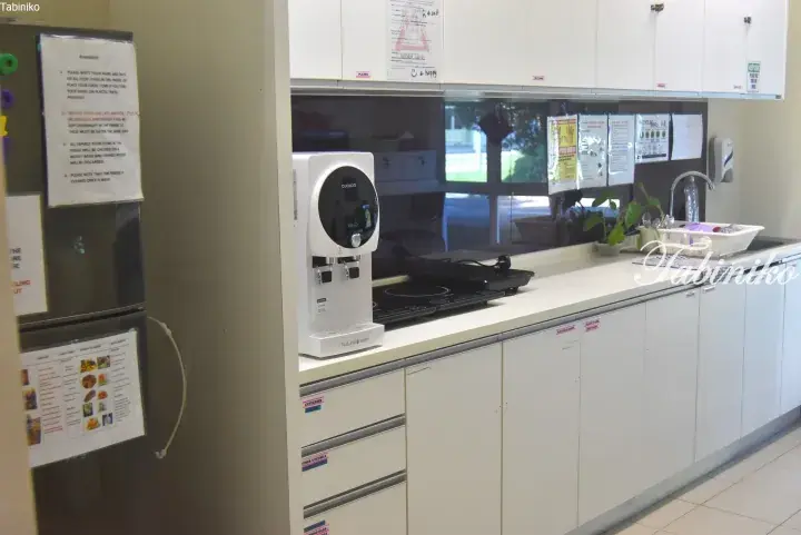 Modern kitchenette with white cabinets, a Cuckoo water dispenser, induction cooktop, and sink in a shared facility.