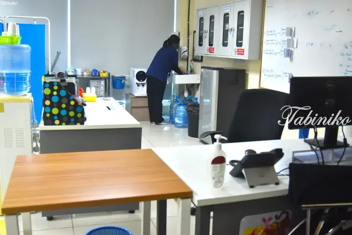 A nurse in blue scrubs washes hands at a sink in a modern clinic reception area with white cabinets and a whiteboard.