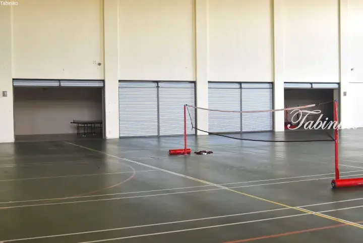Empty indoor sports hall with badminton court markings and portable net, featuring large roller shutter doors.