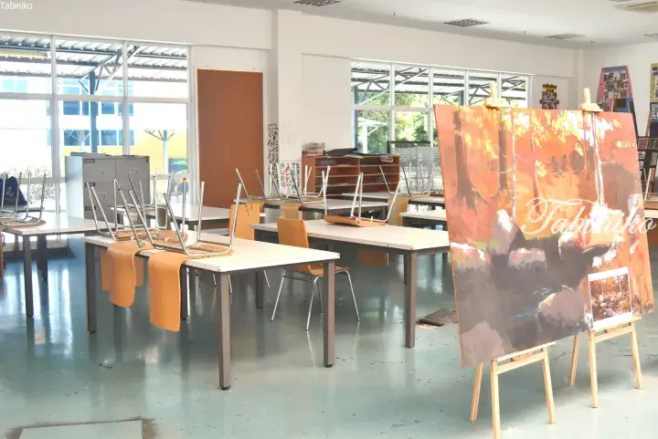 Art classroom with chairs on tables and a large landscape painting on an easel in a bright room.