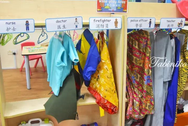 Colorful dress-up costumes hanging in a role-play area with bilingual labels for nurse, doctor, firefighter, police officer, and teacher.