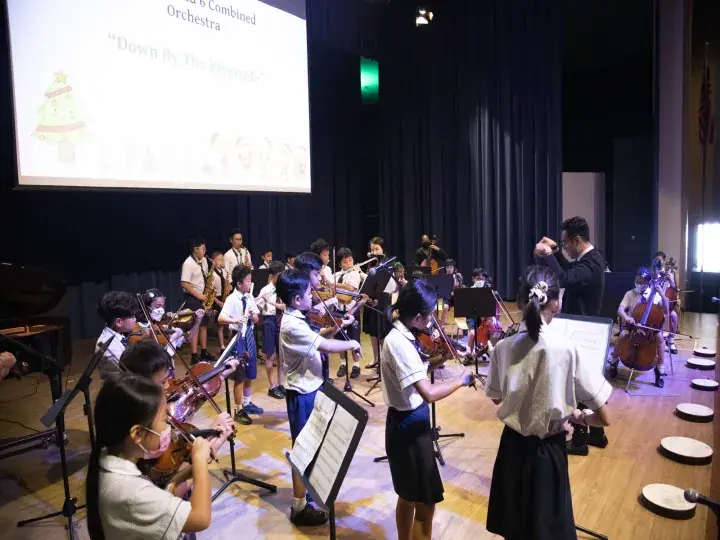 Students perform in a school orchestra concert, playing violins and cellos on stage under a screen showing ‘Down By The Riverside’.
