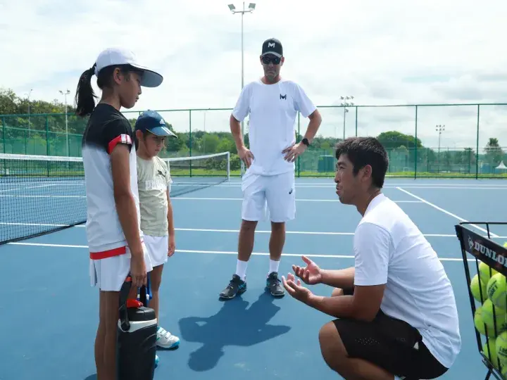 Tennis coach instructs two young students on a blue court while another coach observes nearby.
