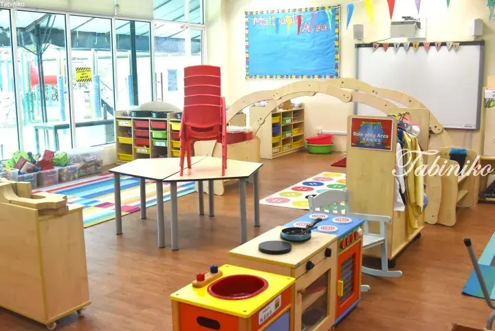 Bright preschool classroom with wooden play kitchen, role-play area, and colorful learning zones.
