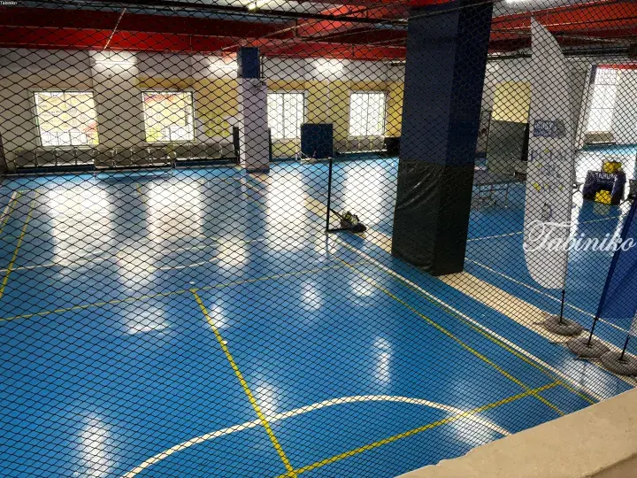 View of an indoor badminton court with blue flooring and yellow lines, seen through a protective safety net.