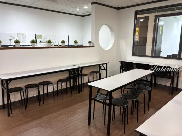 Modern study room with long white tables, black stools, and a kitchen pass-through window in a Malaysia property.