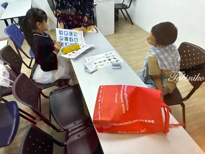Two children sit at a white table with learning worksheets and a red Original Classic shopping bag in a classroom.