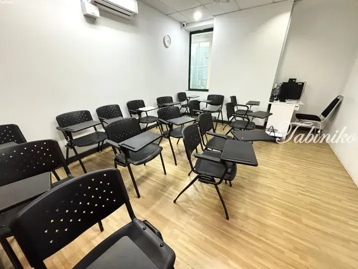 Modern classroom with black student desks and chairs arranged in rows, featuring a teacher’s desk, window, and air conditioning unit.