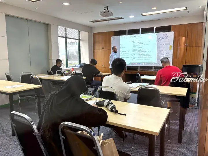 Students in a modern classroom with large windows overlooking a city, attending an English lesson projected on a whiteboard.