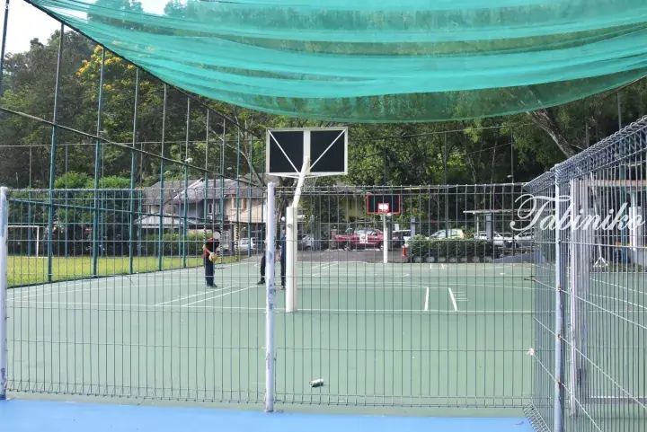 Outdoor basketball court with green flooring and overhead shade net, enclosed by metal fencing in a residential area.
