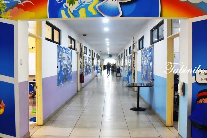 Bright hallway with blue and purple walls, bulletin boards, and a person walking in a Malaysian school or institution.