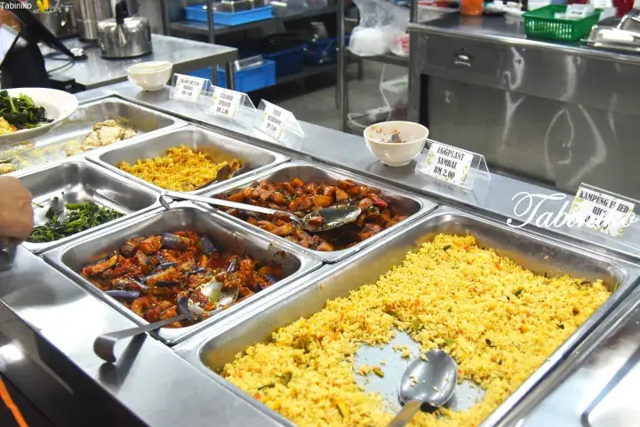 Buffet-style food trays at a Malaysian restaurant featuring Kampung Fried Rice, Eggplant Sambal, and Steamed Spinach.