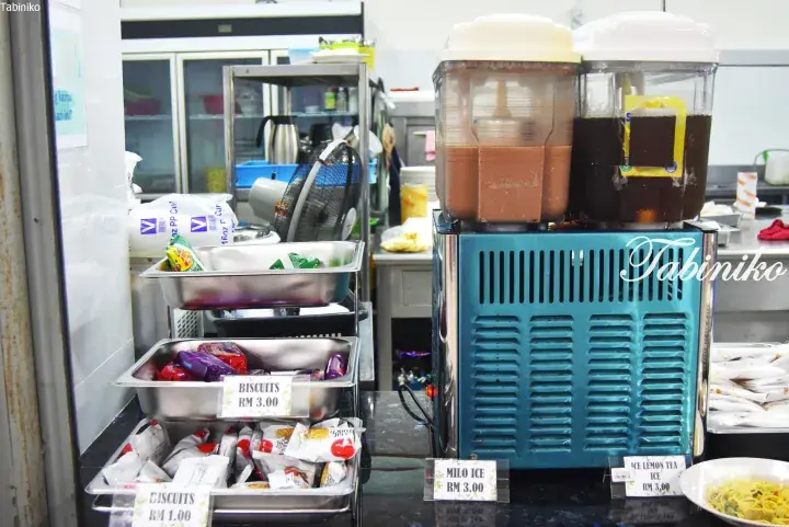 Self-service drink dispensers with Milo Ice and Ice Lemon Tea priced at RM 3.00 alongside trays of biscuits.