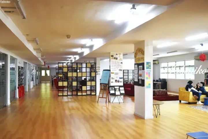 Bright school hallway with wooden floors, glass classrooms, and a lounge area where students relax on yellow sofas.