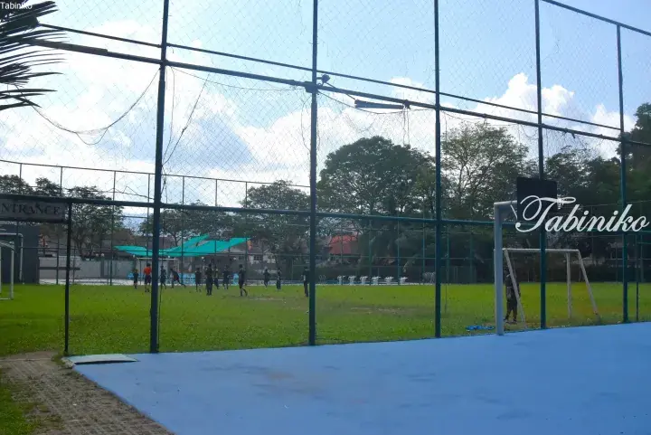 Football players training on a green pitch behind a tall chain-link fence at a sports complex.