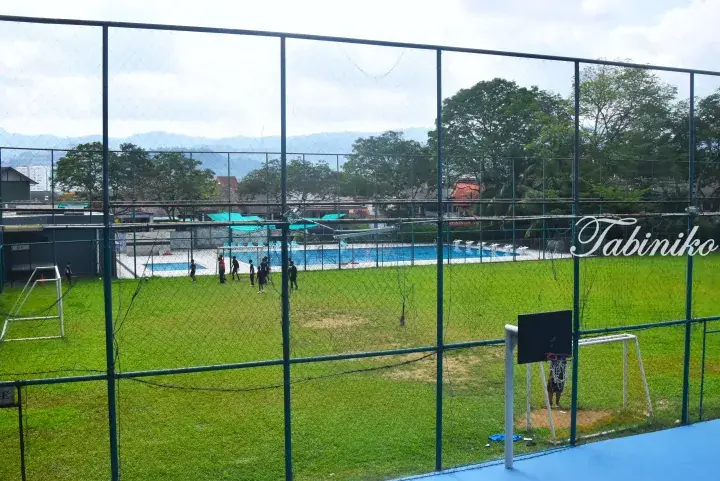 View of a school sports field with a swimming pool, surrounded by a fence with mountains in the background.