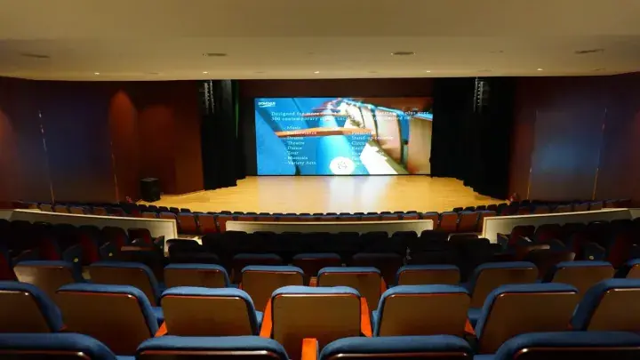 Interior view of a modern auditorium with tiered blue seating facing a wooden stage and large presentation screen.